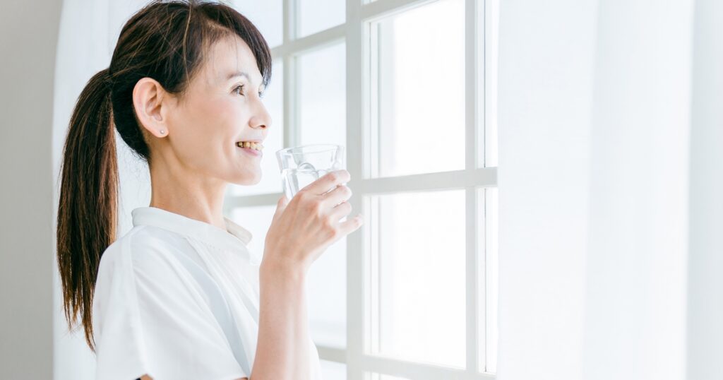 朝に窓を眺めながら、水を飲むミドル女性